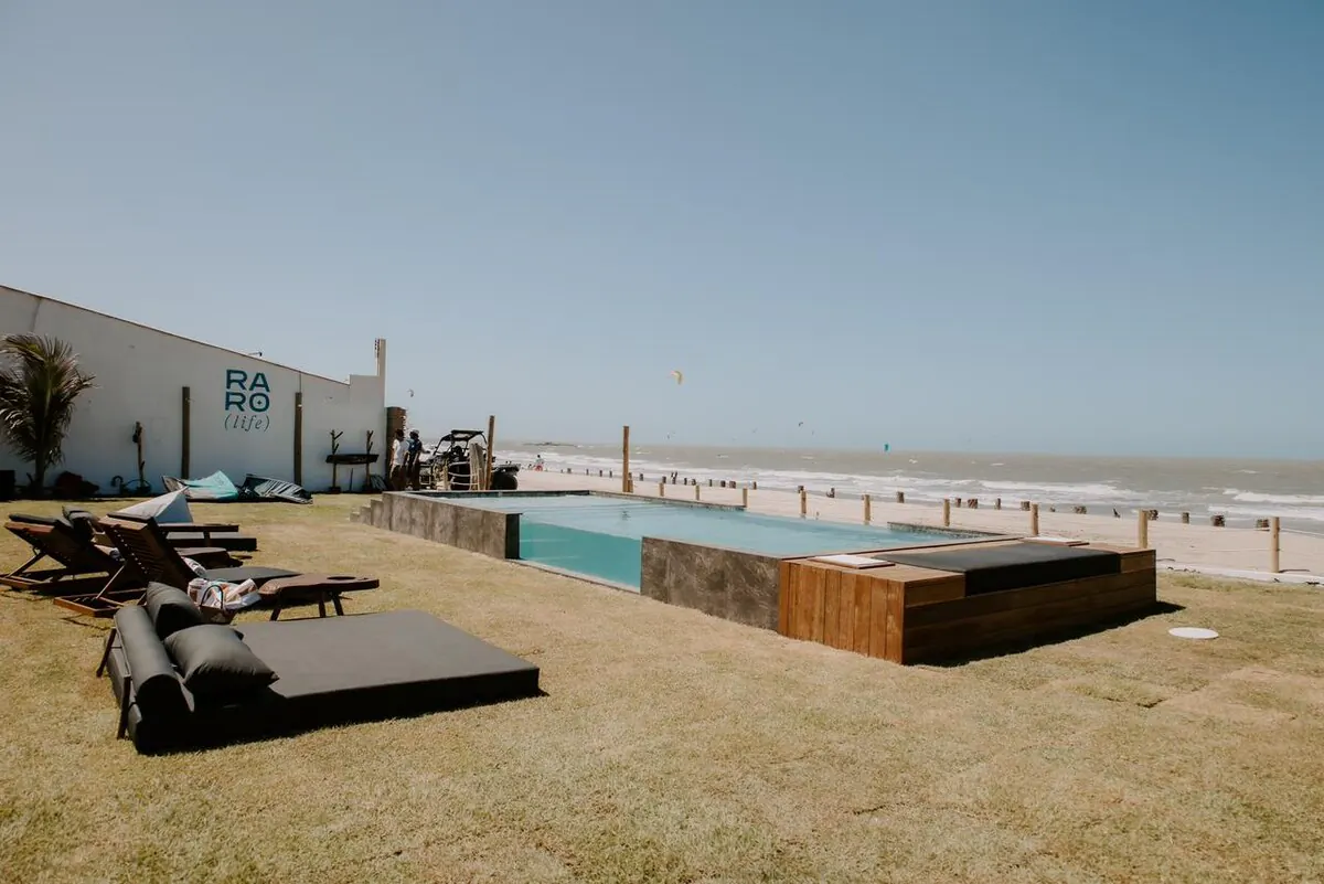 Gramado do beach club com a piscina, o muro com a assinatura RARO (life) e kites no mar